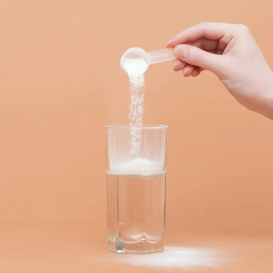 Ein klarer Glasbecher, der mit Wasser gefüllt ist und einen Strom weißen Pulvers aus einem transparenten Plastik-Messlöffel erhält.